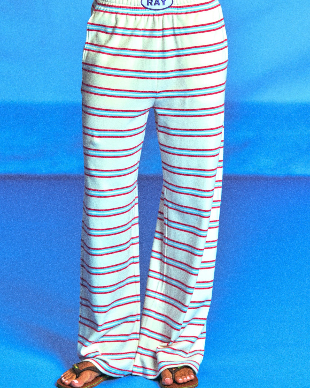 White wide-leg lounge pants with thin red and blue horizontal stripes, featuring an elastic waistband with a blue oval logo that reads 'RAY'. The pants are modeled from the waist down on a bright blue studio background.