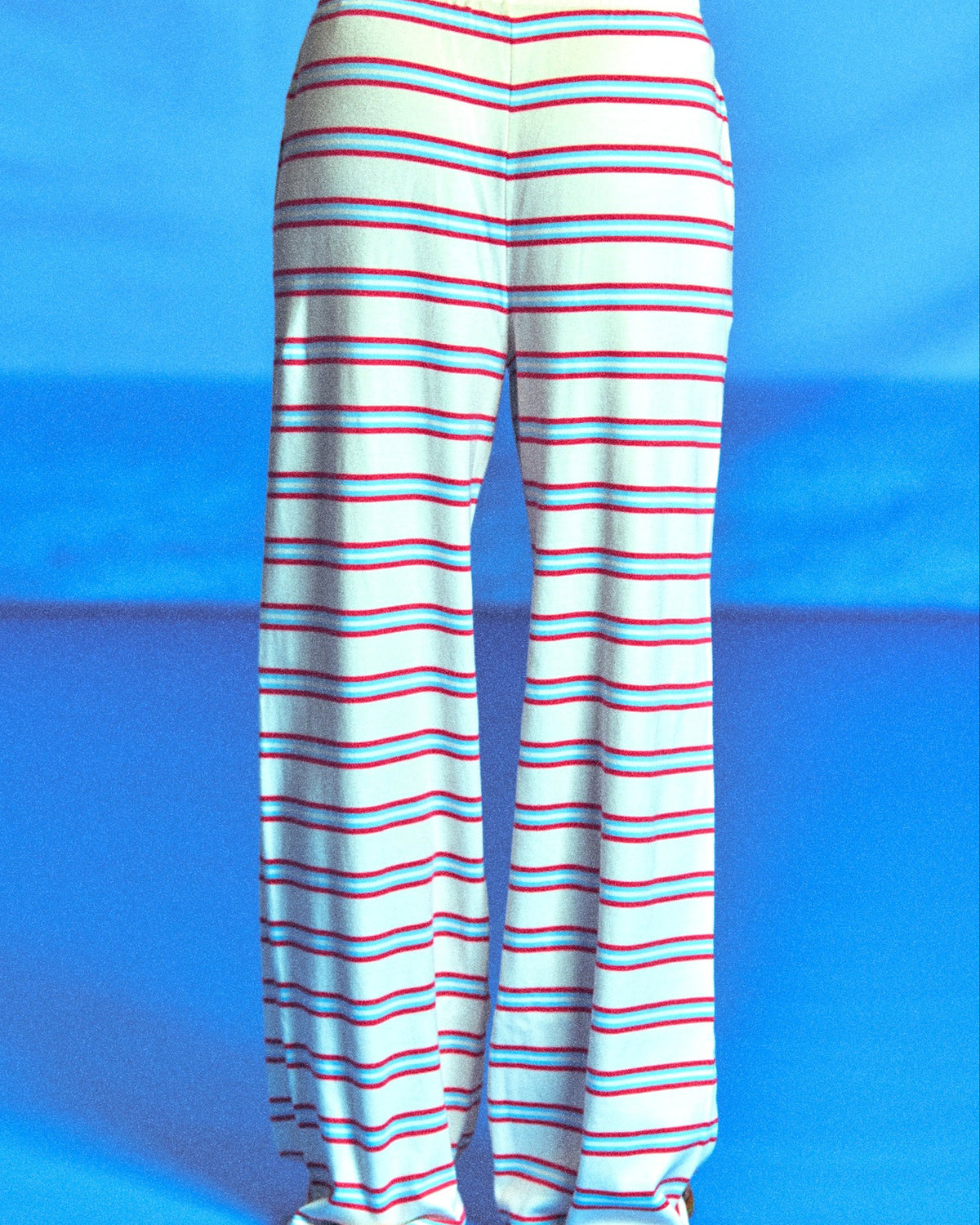 White wide-leg pants with thin red and blue horizontal stripes, shown from the back on a model against a bright blue studio background.