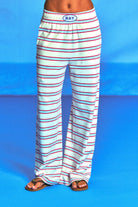 White wide-leg lounge pants with thin red and blue horizontal stripes, featuring an elastic waistband with a blue oval logo that reads 'RAY'. The pants are modeled from the waist down on a bright blue studio background.
