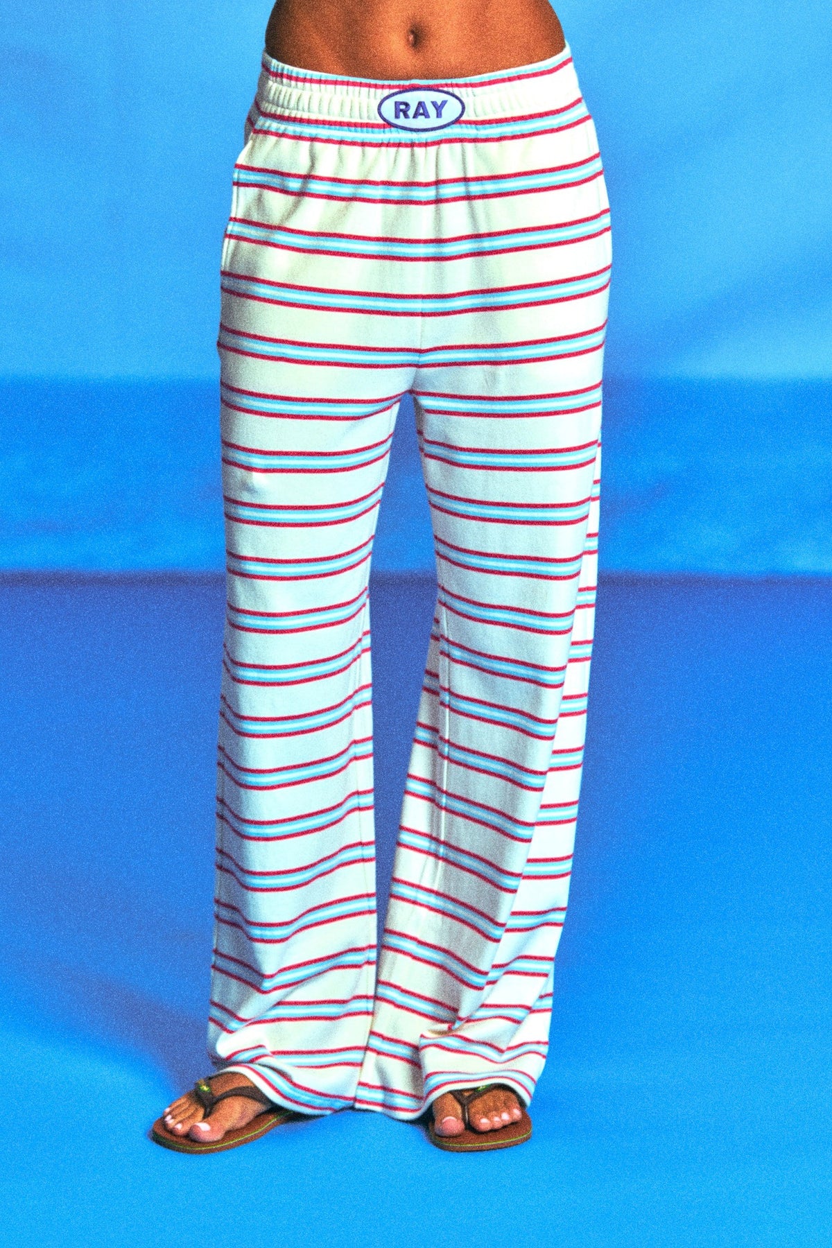 White wide-leg lounge pants with thin red and blue horizontal stripes, featuring an elastic waistband with a blue oval logo that reads 'RAY'. The pants are modeled from the waist down on a bright blue studio background.