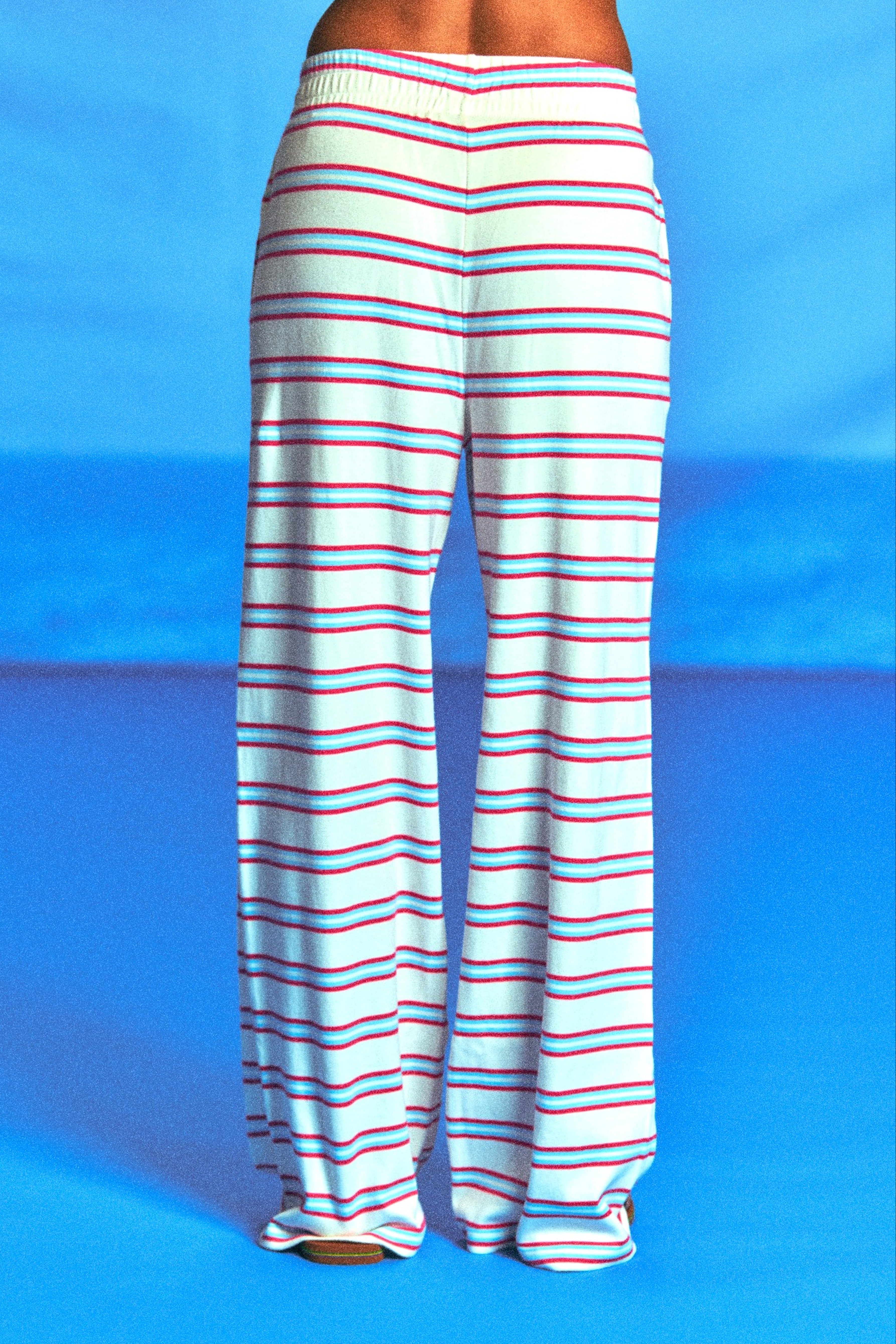 White wide-leg pants with thin red and blue horizontal stripes, shown from the back on a model against a bright blue studio background.
